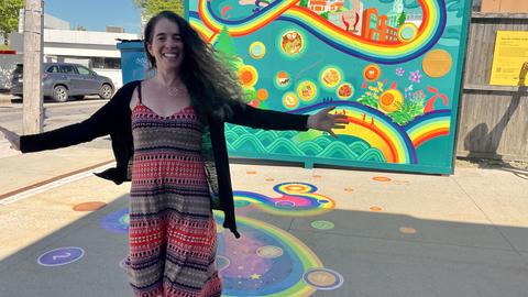Artist stands in front of outdoor mural with arms wide open smiling
