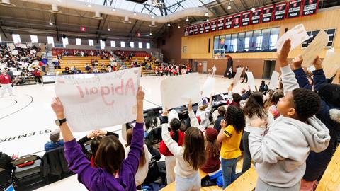 kids In the auditorium stands in the gymnasium cheering on the basketball team holding up signs