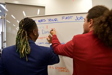 Two people write down their answers on a white board. The question reads: "What was your first job?"