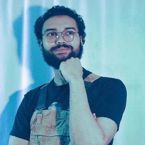 man wearing glasses and overalls thinks pensively for photo with chin sitting on hand