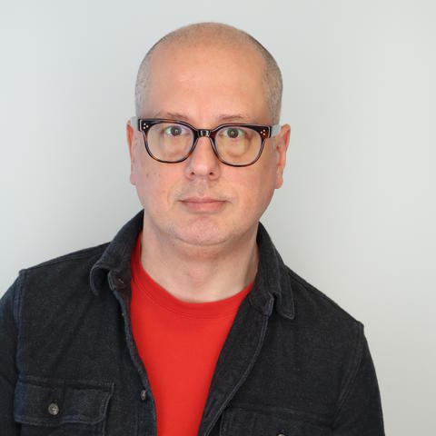 Headshot of artist ansis purins, wearing glasses, red shirt, and black button jacket against white wall
