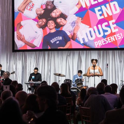 kids playing instruments on stage in front of audience with west end house screen in the background