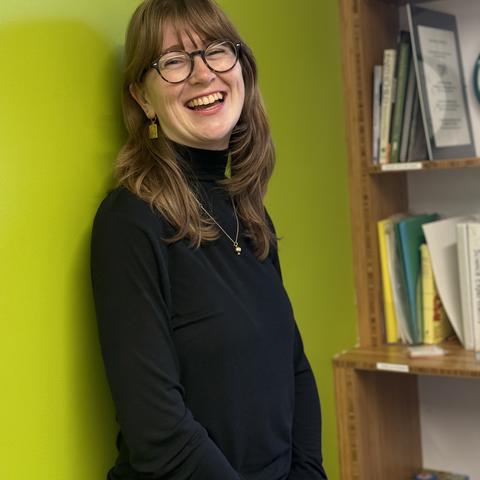 Woman with glasses stands against green wall next to bookshelf smiling