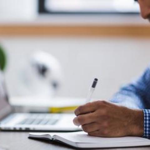 man sitting at desk with lap top writing something with a pen