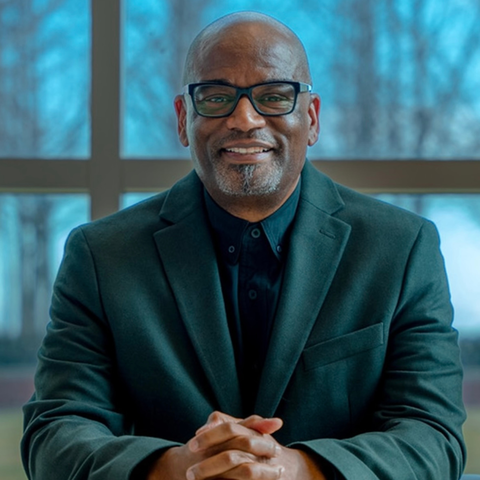Man wearing glasses and suit smiles at camera with folded hands